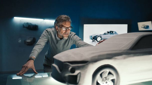 Automobil-Ingenieur berührt das Modell des Prototyp-Autos aus Ton, um letzte Änderungen am Design vorzunehmen. Grauhaariger Auto-Entwickler mit gerahmter Brille in einem Autohersteller. - Foto, Bild