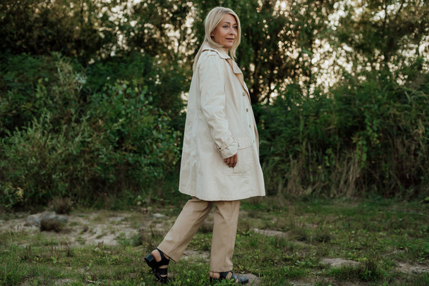 A beautiful adult woman with blond hair dressed in a light raincoat stands still - Foto, Imagen