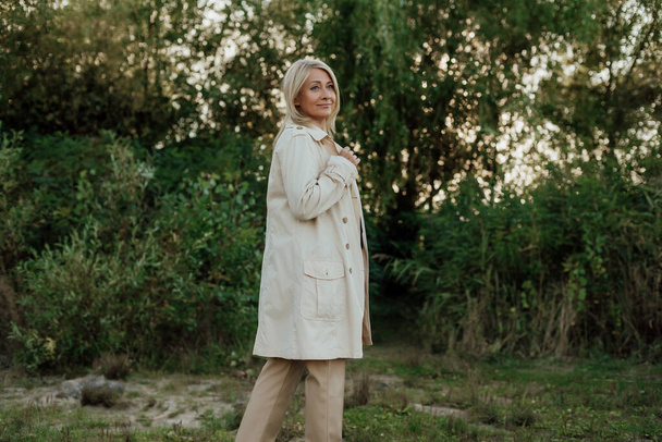 Uma bela mulher adulta com cabelo loiro vestido com uma capa de chuva leve permanece parada - Foto, Imagem