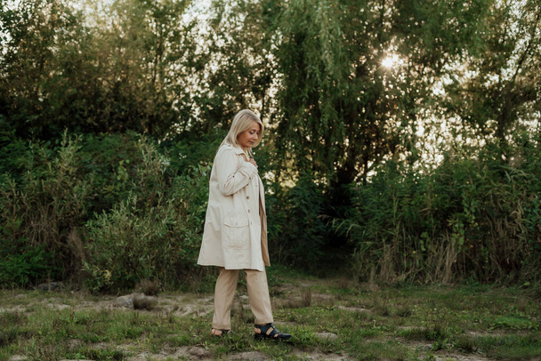 Piękna dorosła kobieta z blond włosami ubrana w jasną płaszcz przeciwdeszczowy stoi nieruchomo - Zdjęcie, obraz