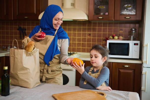 Nahöstliche Muslimin im Hijab, legt eine Tüte mit Lebensmitteln auf den Tisch. Das kleine Mädchen hilft ihrer liebevollen Mutter beim Auspacken von Einkaufstüten mit gesundem Essen in der Küche. Umweltfreundliche Papierverpackungen - Foto, Bild