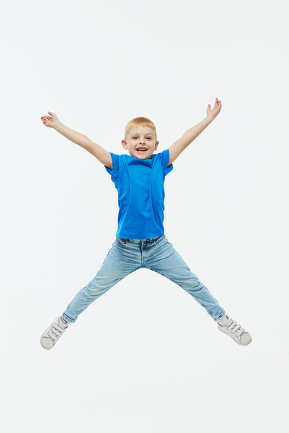 Stylish child boy model in a blue casual t-shirt joyfully jumping high on a white isolated background - Photo, image