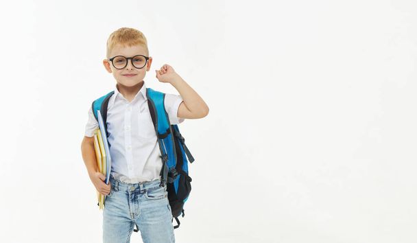 Back to school. Funny little boy from elementary school with a book, a backpack and glasses on a white background. Laughs merrily. Preparation for school. Concept of early development - Foto, imagen