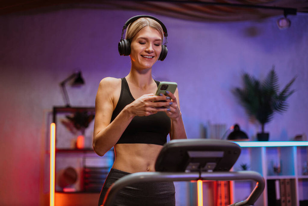 Low angle view of beautiful caucasian woman listening favorite music on phone, exercising, having workout on a treadmill in living room. Happy female running, having cardio training at evening time. - 写真・画像