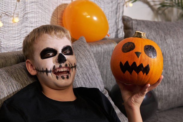 Ένα τρομακτικό παιδί με μακιγιάζ σε σχήμα σκελετού και με μια κολοκύθα στα χέρια του γελάει κοιτώντας την κάμερα. Το παιδί γιορτάζει Απόκριες - Φωτογραφία, εικόνα