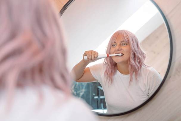 Blanke roze haar vrouw poetsen tanden met elektrische tandenborstel in de badkamer. Moderne huiselijke levensstijl. Hygiëne en tandheelkundige verzorging - Foto, afbeelding