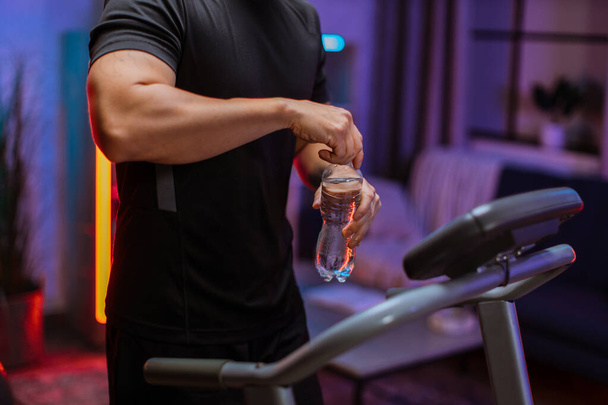 Close up of hands of sports man drinking opening water bottle, doing fitness exercise, running on treadmill . Athletic and muscular male having workout indoor, at modern apartment at night time. - 写真・画像