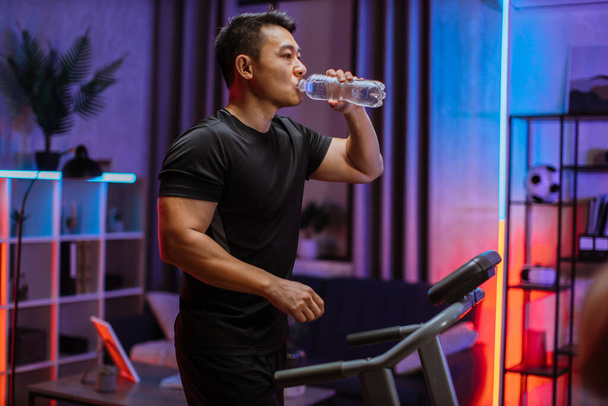Привлекательный молодой спортивный азиат, пьющий воду из бутылки, занимающийся фитнесом, бегущий по беговой дорожке. Спортивный и мускулистый мужчина, тренирующийся в помещении, в современной квартире в ночное время дома. - Фото, изображение