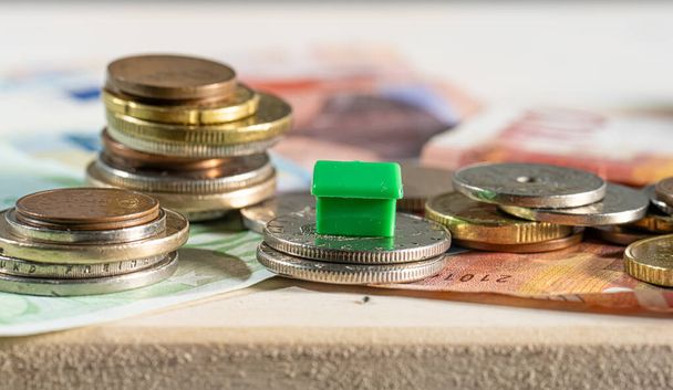 Mały domek pomiędzy stosami monet reprezentujących gospodarkę mieszkaniową oraz koszty kredytu mieszkaniowego i odsetki. - Zdjęcie, obraz