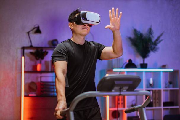 Portrait of young active sporty asian man in black sportswear training at home, doing cardio exercise on treadmill using vr glasses at evening time. - Fotografie, Obrázek
