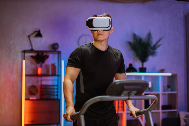 Attractive young sports asian man doing fitness exercise, running on treadmill using virtual reality glasses. Athletic and muscular male having actively workout indoor at night time. - Foto, afbeelding