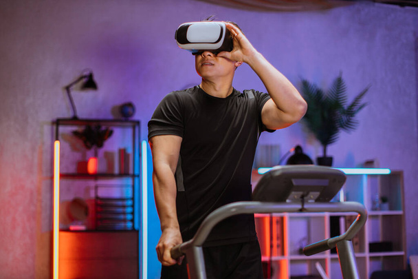 Attractive young sports asian man doing fitness exercise, running on treadmill using virtual reality glasses. Athletic and muscular male having actively workout indoor at night time. - Fotó, kép