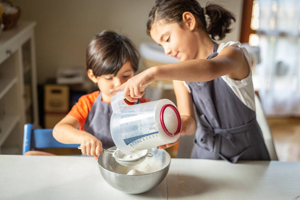 dos niñas con delantales grises preparándose para hornear y tamizar la harina en un tazón de metal - Foto, Imagen