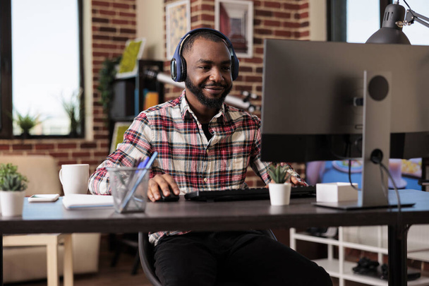 Büroangestellte arbeiten von zu Hause aus am Computer und hören Musik oder Podcasts online über Kopfhörer. Mann benutzt Headset und surft Internet-Website und erledigt freiberufliche Arbeit. - Foto, Bild