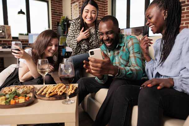 Multi ethnic bunch young adults guffawing at african american man smart phone during friends house party, drinking wine, smiling, laughing, happily enjoying mates gathering. - Photo, Image