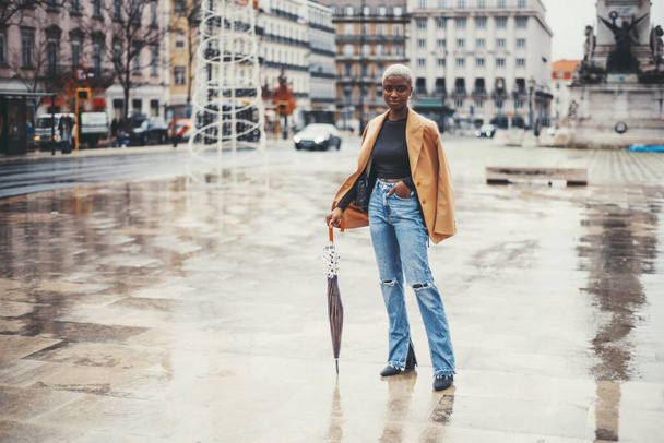 A young svelte black female with short hair painted white is standing on the wet ground of Lisbon city center after the rain and leaning on her umbrella, with a copy space area on the left - Zdjęcie, obraz
