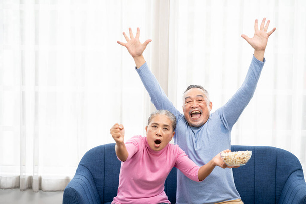 Взрослая пара, пожилые мужчины и женщины смотрят телевизор, старшие спортивные болельщики празднуют любимую командную победу, сидят на уютном диване и едят попкорн закуски дома, наслаждаясь выходными. Счастливые пенсионеры. - Фото, изображение