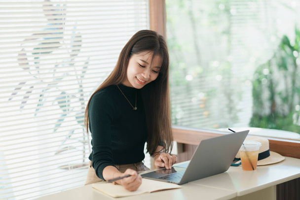 Asiatische Freiberuflerin lächelt, tippt auf Tastatur und arbeitet zu Hause auf einem Laptop auf einem Holztisch. Unternehmerin, die für ihr Geschäft im Café arbeitet. Geschäftliches Arbeiten an jedem Ort. - Foto, Bild