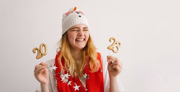 Hauska nuori tyttö uudenvuoden hattu ja punainen huivi pitää numerot 2023 käsissään. Uusi vuosi ja joulun käsite. - Valokuva, kuva
