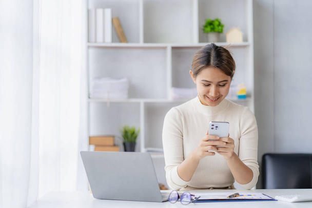 Exitosa empresaria asiática sonriendo con un smartphone hablando con un cliente en su escritorio en la oficina de una compañía de concepto de contabilidad financiera. - Foto, imagen