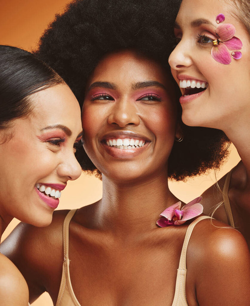 Women, beauty and flowers, makeup and diversity of models on orange studio background. Skincare, face and happy, young and elegant female group posing together with cosmetics, orchids or pink plants - Fotó, kép