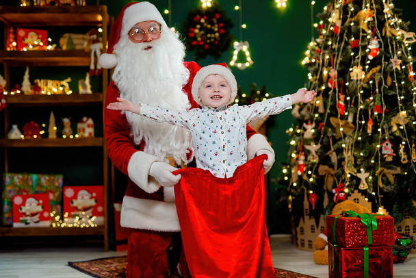 Happy little boy having fun with santa claus sitting in a sack with gifts near Christmas tree. New Year concept. - Photo, image