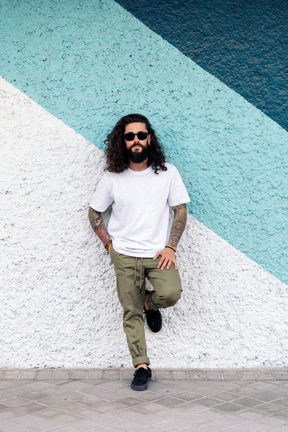 Cool hipster man with long hair posing and looking at camera against blue background. - Fotó, kép