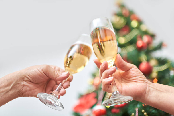 Unknown adult couple toasting during Christmas or New Year, celebrating holidays together. Low angle view of a female and male hand clinking a pair of glasses, with a decorated tree in the background - Photo, Image