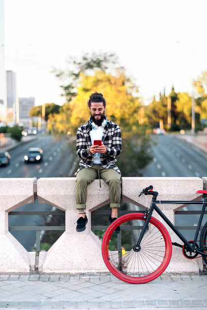 Cool man with big beard sitting in the city next to his bike and using his mobile phone. - Photo, Image