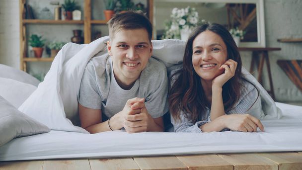 Portrait of happy couple lying in bed under blanket looking at camera laughing and smiling. Loving married people and happiness concept. - Photo, image