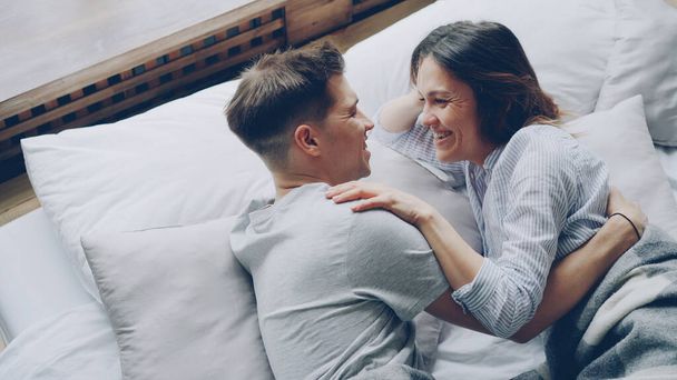 Attractive asian woman is lying in bed with her boyfriend touching him while he is hugging her, they are talking and laughing. Tenderness and love concept. - 写真・画像