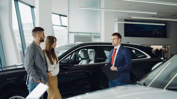 Car dealership worker is talking to beautiful couple young bearded man and his attractive wife while standing near luxury car in auto salon. Choosing new automobile concept. - Фото, изображение