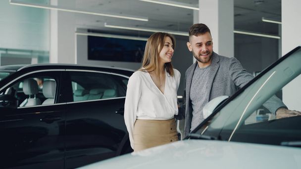 Attractive young couple confident bearded guy and his smiling pretty girlfriend are choosing new automobile together looking at luxury car in auto dealership and talking. - Valokuva, kuva