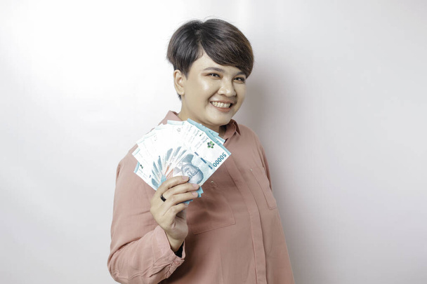 A happy young woman is wearing pink shirt and holding cash money in Indonesian rupiah isolated by white background - Zdjęcie, obraz