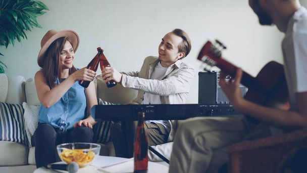Cheerful young friends are having fun talking, singing and drinking beer during rehearsal. Guitarist is playing, woman and man are clanging bottles and laughing. - Photo, Image