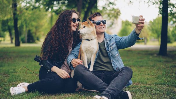 Handsome young man is taking selfie with his pretty wife and cute dog, all wearing sunglasses. Guy is holding smartphone taking funny pictures and posing. - Foto, afbeelding