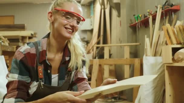 Carpenter beautiful girl wearing safety glasses. Choose long slats of sawn timber to make furniture with care, dusting, cleaning and wanting to use this lumber with smile. - Footage, Video