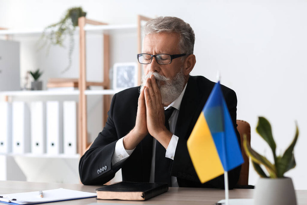 Mature man with flag of Ukraine praying in office - Foto, Imagen