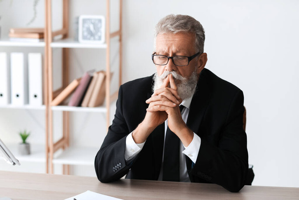 Зрелый бизнесмен молится за столом в офисе - Фото, изображение