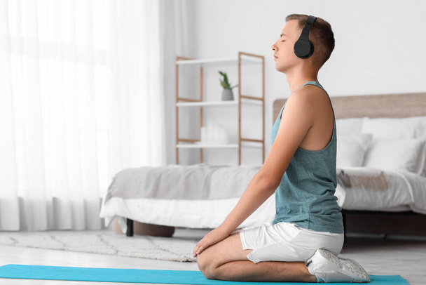 Junger Mann mit Kopfhörern meditiert auf Matte im Schlafzimmer - Foto, Bild
