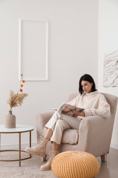 Молодая женщина читает журнал в бежевом кресле дома - Фото, изображение