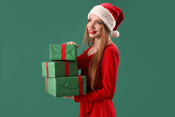 Beautiful young woman in Santa hat with Christmas gifts on green background - Фото, изображение