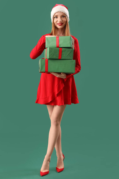 Beautiful young woman in Santa hat with Christmas gifts on green background - 写真・画像