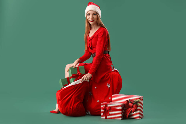 Beautiful young woman in Santa hat with bag of gifts sitting on green background - Photo, Image