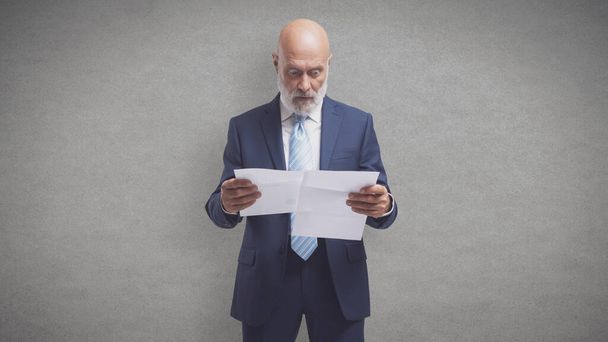 Shocked mature businessman holding expensive utility bills and invoices: energy prices, crisis and costs concept  - Photo, image