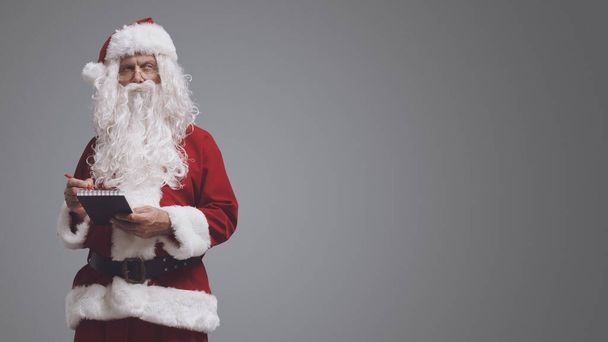 Pensive Santa Claus γράφοντας σημειώσεις και τη λίστα επιθυμιών σε ένα σημειωματάριο, Χριστούγεννα και διακοπές έννοια - Φωτογραφία, εικόνα