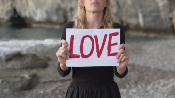 Close-up, a woman standing by the sea with a sheet of paper with the inscription LOVE - Footage, Video