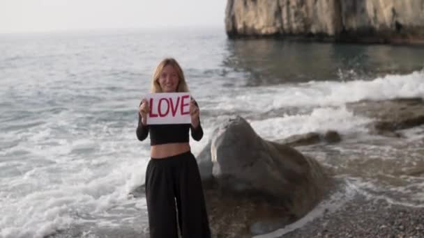 A happy woman stands by the sea and holds in her hands a paper with the inscription LOVE. - Footage, Video