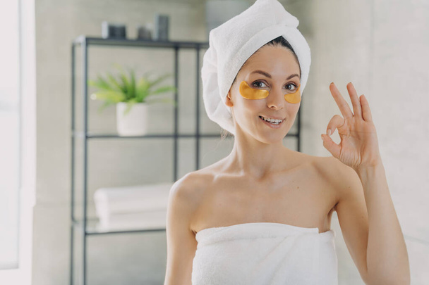 Girl applies collagen eye patches and gestures ok. Attractive woman wrapped in towel after spa procedures. Young hispanic lady does hair and body care. Relaxation at modern spa resort. - Photo, Image