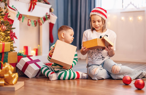 妹と弟のクリスマスツリーの近くの床の上に屋内に座っている。愛の概念、家族関係とお祝いのクリスマスと新年. - 写真・画像